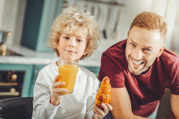 かわいい巻き毛の男の子食べるクロワッサンとオレンジ ジュースを飲む - 写真・画像