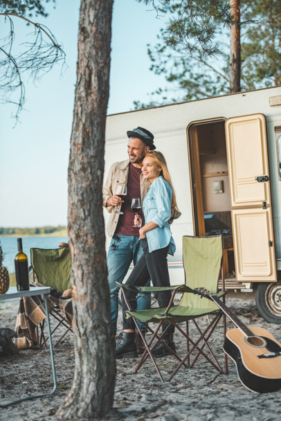 young couple with glasses of wine standing near campervan  - Foto, Bild