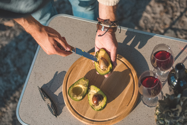 partial view of man cutting avocado on wooden board on table with wineglasses and sunglasses - Photo, Image