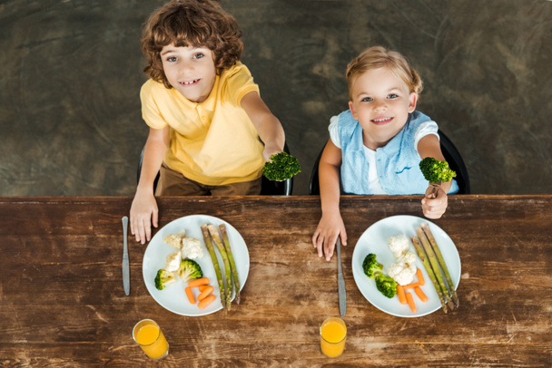 high angle view of cute happy children holding forks with broccoli and smiling at camera - Фото, изображение