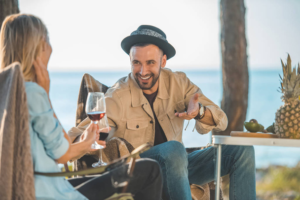 pareja sonriente con copas de vino hablando durante un picnic cerca del mar
 - Foto, imagen