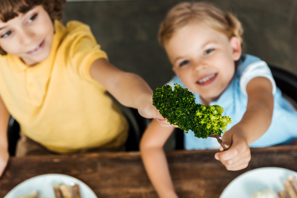 vista de ángulo alto de adorables niños felices sosteniendo horquillas con brócoli
 - Foto, Imagen