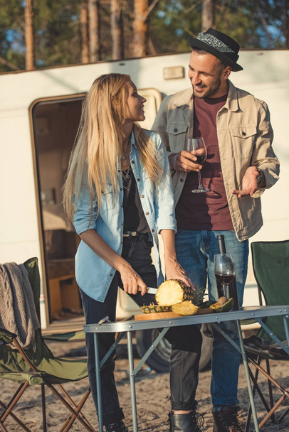 beautiful girl cutting pineapple and looking man with wine near campervan - Fotoğraf, Görsel