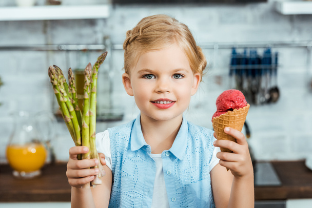 adorable niño feliz sosteniendo delicioso cono de helado y espárragos saludables, sonriendo a la cámara
 - Foto, Imagen