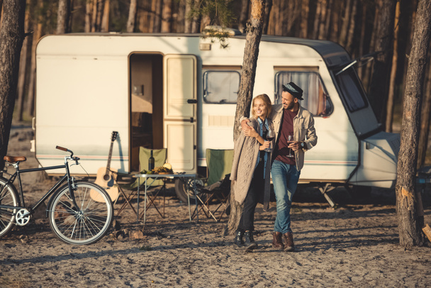 happy couple with glasses of wine walking near trailer in camp - Фото, изображение