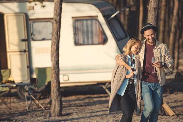 beautiful couple with glasses of wine hugging and walking near campervan - Foto, Imagen