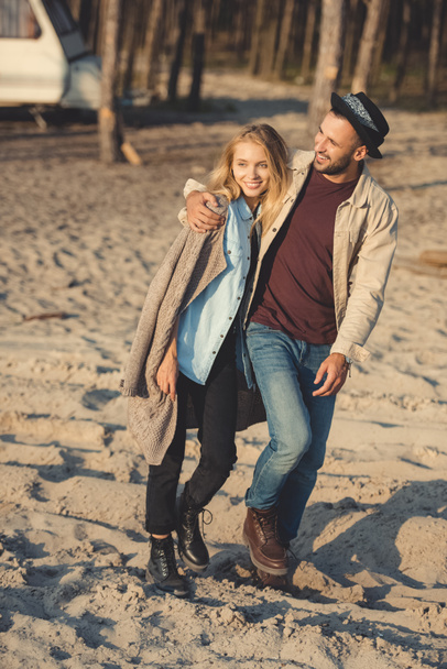 hombre sonriente y mujer atractiva abrazando y caminando en la playa de arena
 - Foto, Imagen
