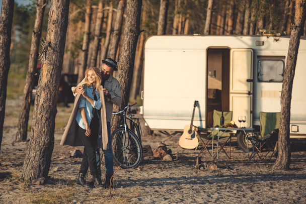 νεαρό ζευγάρι ευτυχισμένο Αγκαλιάζοντας στο στρατόπεδο με ρυμουλκούμενο, ποδήλατο και κιθάρα - Φωτογραφία, εικόνα