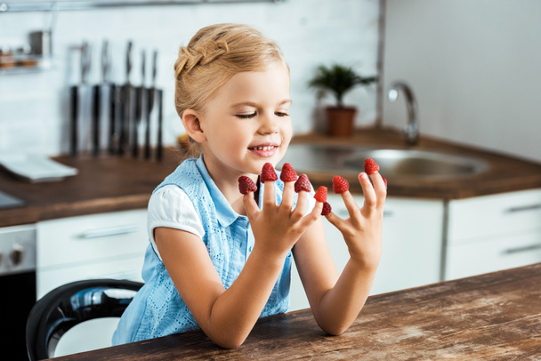 cute happy kid sitting at table and looking at ripe fresh raspberries on fingers  - Zdjęcie, obraz