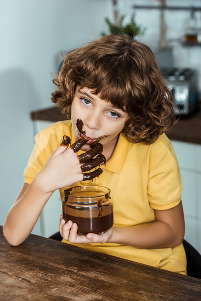 cute little boy eating sweet chocolate spread from glass jar and looking at camera - Foto, Bild