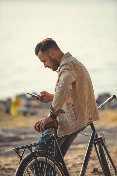 jovem homem bonito usando smartphone e inclinando-se na bicicleta ao ar livre
 - Foto, Imagem
