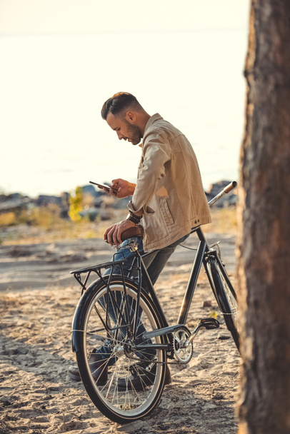 young man using gadget and leaning on bicycle near sea - Φωτογραφία, εικόνα