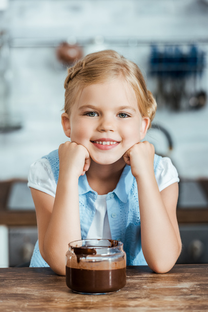 lindo niño feliz sentado en la mesa con chocolate extendido en frasco y sonriendo a la cámara
 - Foto, Imagen
