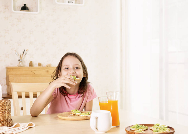 Cute smiling preteen girl having healthy breakfast: avocado sandwich and orange juice. Healthy lifestyle concept, vegetarian food. Superfoods. Living room interior in background - Photo, Image