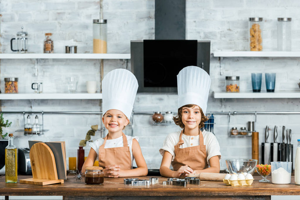 adorable happy kids in aprons and chef hats smiling at camera while cooking together in kitchen - Fotó, kép