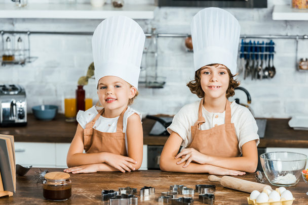 cute happy children in aprons and chef hats smiling at camera while cooking together in kitchen - Foto, immagini