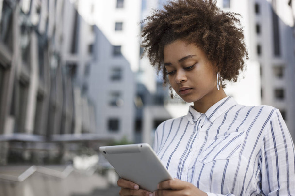 Afro fille américaine est debout dans la rue près de l'immeuble de bureaux et tient un ordinateur portable dans ses mains. Elle travaille sur une tablette.
. - Photo, image