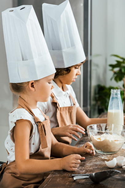 side view of adorable kids in chef hats and aprons preparing dough for cookies in kitchen - Photo, Image