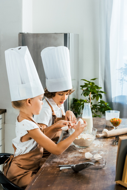 sivukuva söpöistä pienistä lapsista kokin hatuissa ja esiliinoissa valmistamassa taikinaa evästeisiin yhdessä
 - Valokuva, kuva