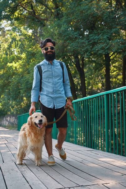 Ochtend wandeling met hond. Jonge man met zijn labrador retriever op de houten brug met bomen en de zon licht in de achtergrond. - Foto, afbeelding