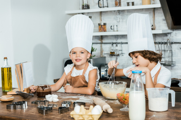 lindos niños pequeños en delantales y sombreros de chef preparando masa para galletas y sonriendo a la cámara
 - Foto, Imagen