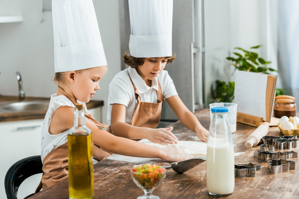 χαριτωμένο μικρό παιδιά σε ποδιές και καπέλα σεφ προετοιμάζει ζύμη για νόστιμο cookies μαζί  - Φωτογραφία, εικόνα