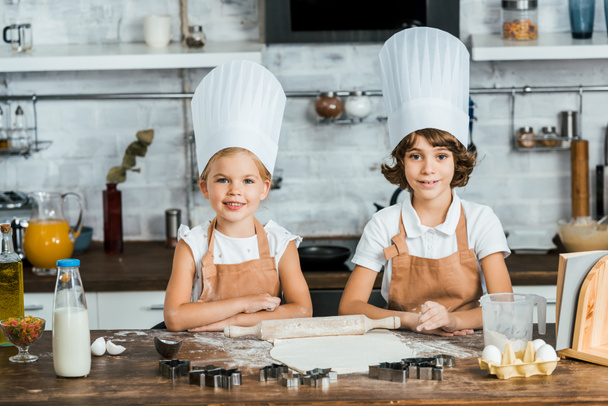 ładny szczęśliwe dzieci w kapeluszach chef przygotowania ciasta ciasteczka i uśmiecha się do kamery  - Zdjęcie, obraz