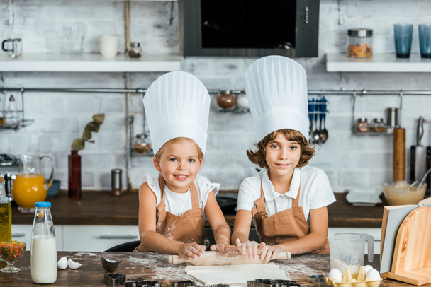 adorable happy kids in chef hats preparing dough for cookies and smiling at camera  - Φωτογραφία, εικόνα