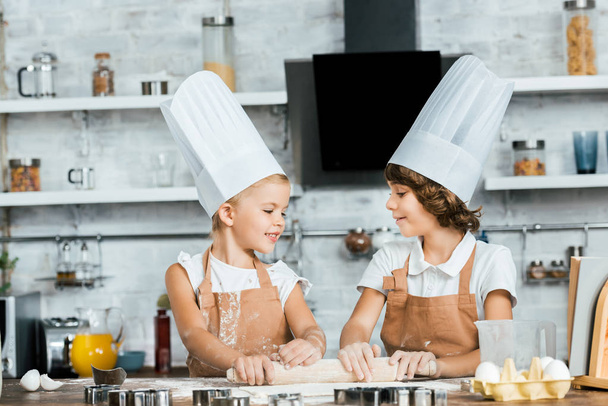 Urocza szczęśliwe dzieci w kapeluszach chef przygotowania ciasta ciasteczka i uśmiechający się wzajemnie - Zdjęcie, obraz