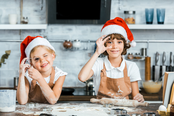 αξιολάτρευτα πιτσιρίκια σε καπέλα santa προετοιμασία χριστουγεννιάτικα μπισκότα και να χαμογελά στη φωτογραφική μηχανή  - Φωτογραφία, εικόνα