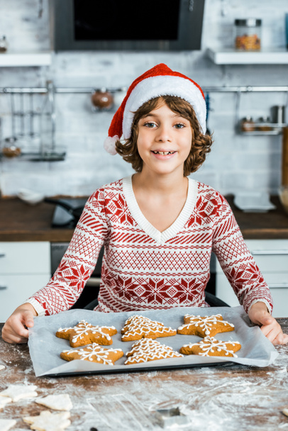 lindo feliz chico en santa hat celebración de bandeja para hornear con galletas de jengibre y sonriendo a la cámara
  - Foto, imagen
