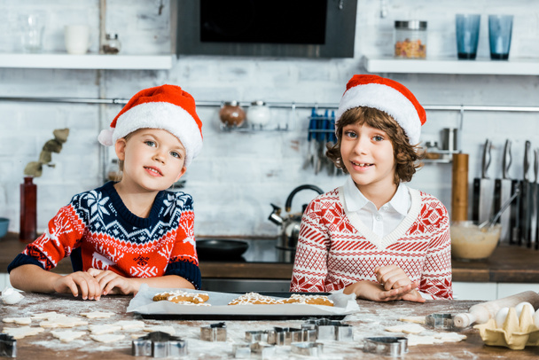 carino bambini felici in cappelli di Babbo Natale preparare biscotti allo zenzero e sorridente alla fotocamera
 - Foto, immagini