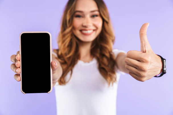 Close up of an excited young girl standing isolated over violet background, showing blank screen mobile phone, showing thumbs up - Valokuva, kuva