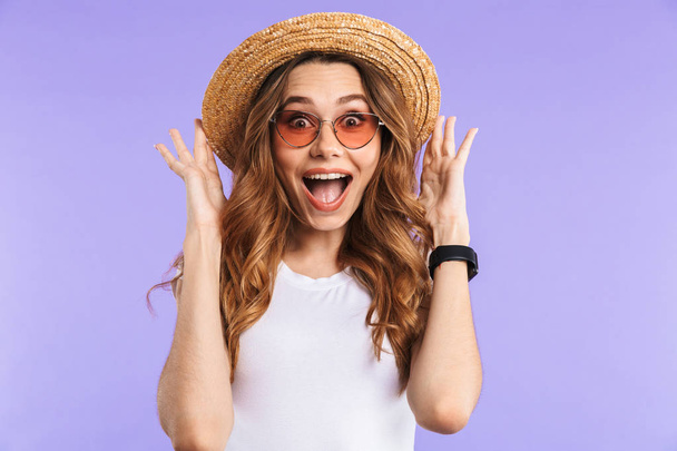 Portrait of a cheerful young girl in sunglasses and straw hat isolated over violet background, looking at camera - Fotó, kép
