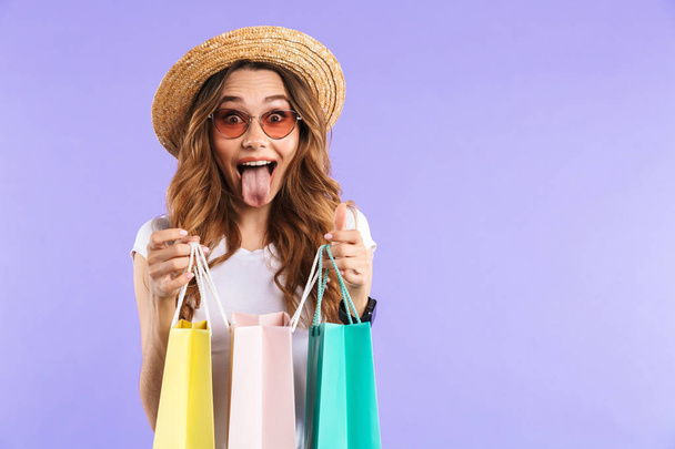 Photo of excited surprised cute woman isolated over purple wall background holding shopping bags showing tongue. - 写真・画像