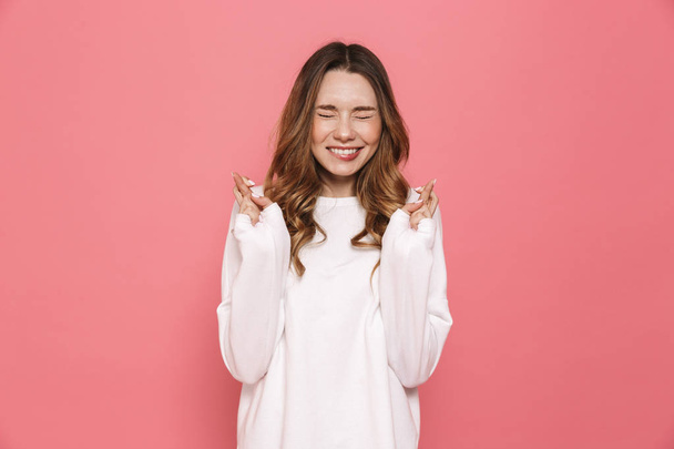 Portrait of a worried young casual girl holding fingers crossed for good luck isolated over pink background - Photo, image