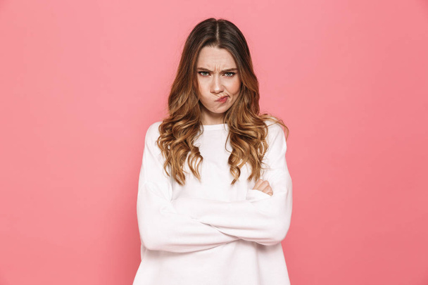 Portrait of an upset young casual girl standing with arms folded isolated over pink background - 写真・画像