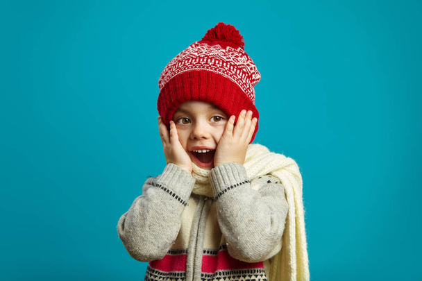 Surprised child girl in winter hat on blue isolated background, put her hands to cheek, opened mouth wide, expresses surprise or shock, received a welcome gift and surprise, joyful child emotional - Photo, Image