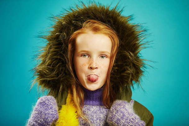 Funny child girl in winter jacket with hood showing tongue on blue isolated background. - Φωτογραφία, εικόνα