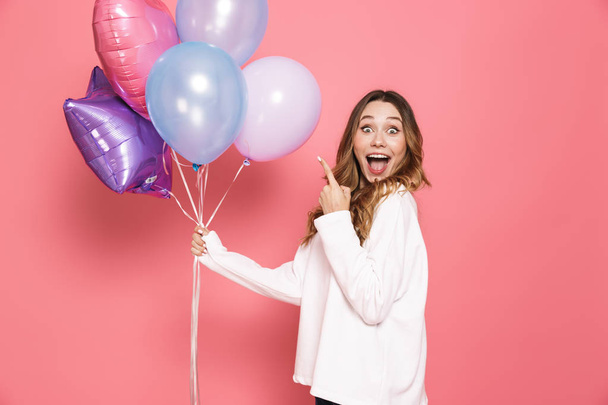 Portrait of a joyful young woman holding bunch of air balloons isolated over pink background, pointing finger - Photo, image