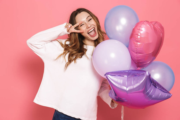 Portrait of a joyful young woman holding bunch of air balloons isolated over pink background, showing peace - Fotografie, Obrázek