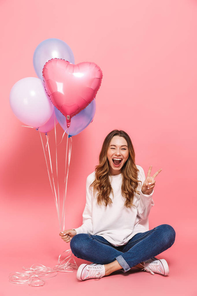 Full length portrait of an excited young woman holding bunch of air balloons while sitting isolated over pink background - Foto, Imagem