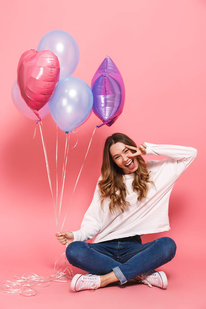 Full length portrait of a cheerful young woman holding bunch of air balloons while sitting isolated over pink background - Foto, afbeelding
