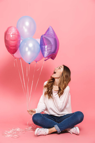 Full length portrait of a lovely young woman holding bunch of air balloons while sitting isolated over pink background - Fotoğraf, Görsel