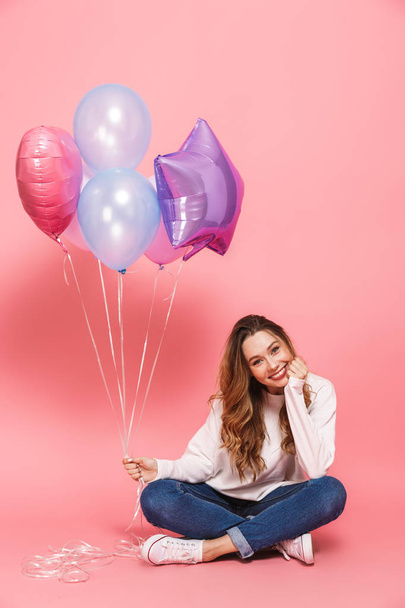 Full length portrait of a smiling young woman holding bunch of air balloons while sitting isolated over pink background - Valokuva, kuva