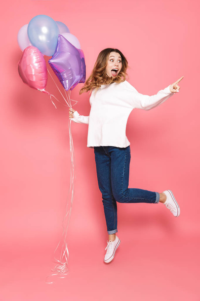 Full length portrait of a cheerful young woman holding bunch of air balloons while jumping isolated over pink background, pointing away - Фото, зображення