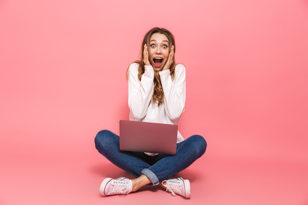 Portrait of a shocked young woman sitting with legs crossed, using laptop computer isolated over pink background - Foto, Imagem