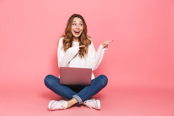 Portrait of a happy young woman sitting with legs crossed, using laptop computer isolated over pink background, pointing away - Foto, afbeelding