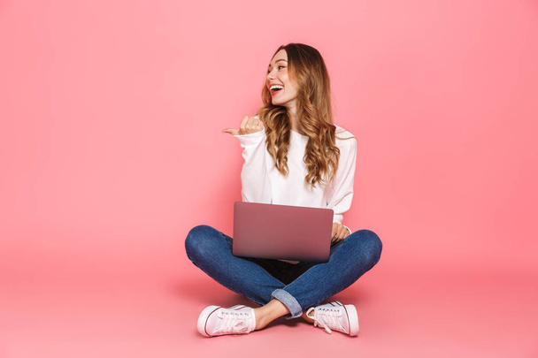 Portrait of a joyful young woman sitting with legs crossed, using laptop computer isolated over pink background, pointing away - Фото, зображення