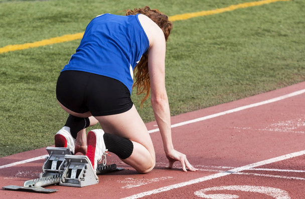 Uma velocista feminina do ensino médio está nos blocos iniciais pronta para iniciar sua corrida de sprint em uma pista vermelha ao ar livre
. - Foto, Imagem